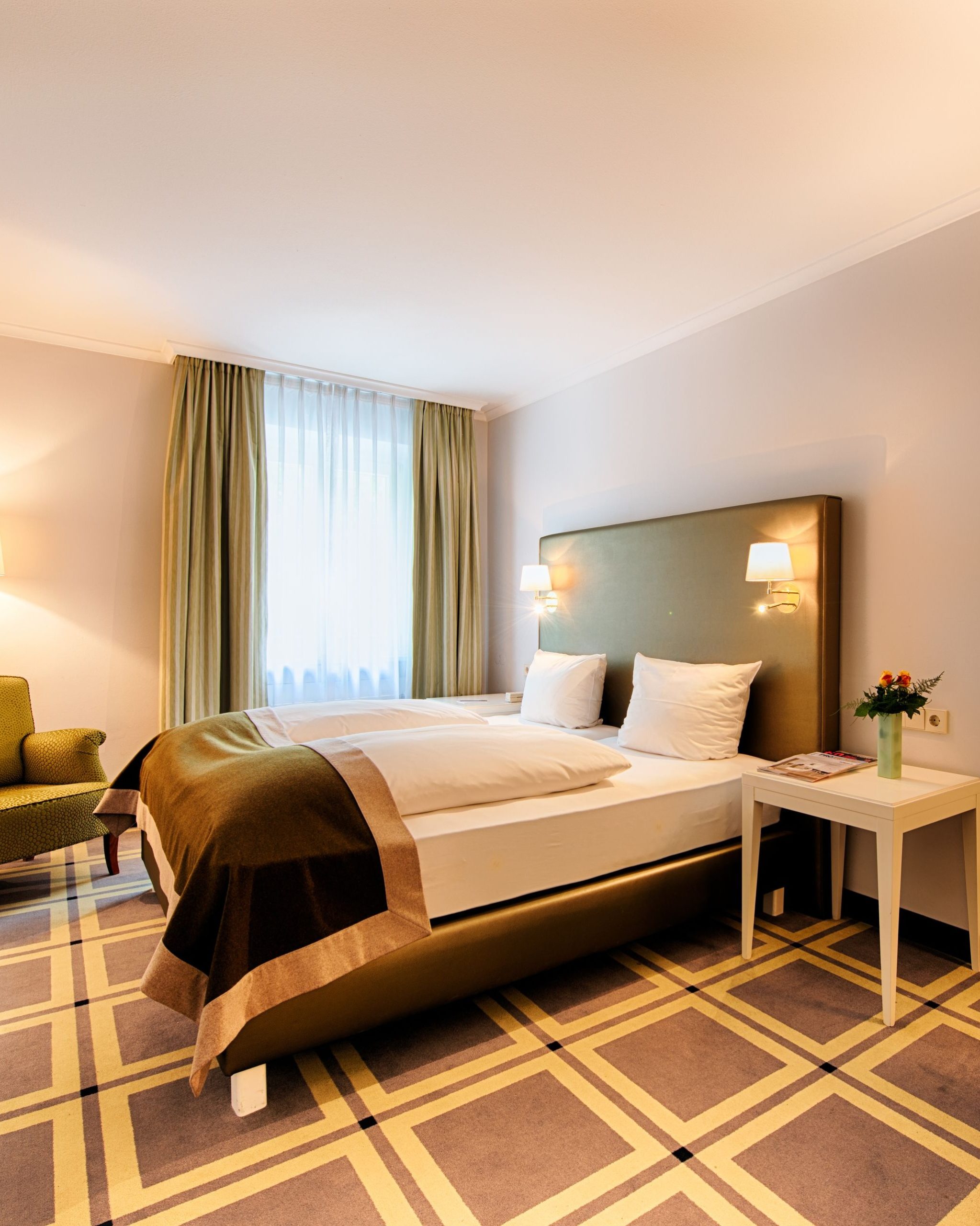 Aufnahme eines Hotelzimmers mit Fokus auf das Bett, Fenster und einem Stuhl in der Ecke.
